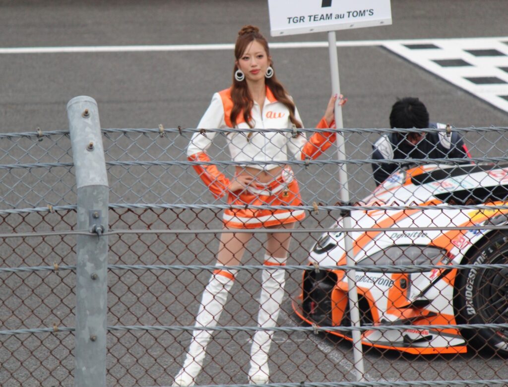 SUPER GTもてぎ2025のスタート前グリッドでポーズをとるTGR TEAM au TOM’Sのアンバサダー。チームカラーのオレンジが映える。